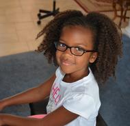 A young girl with glasses and curly hair smiling indoors.
