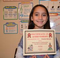 Young girl proudly holding a certificate of achievement in a classroom.