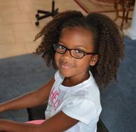 Smiling young girl with glasses and curly hair sitting indoors.