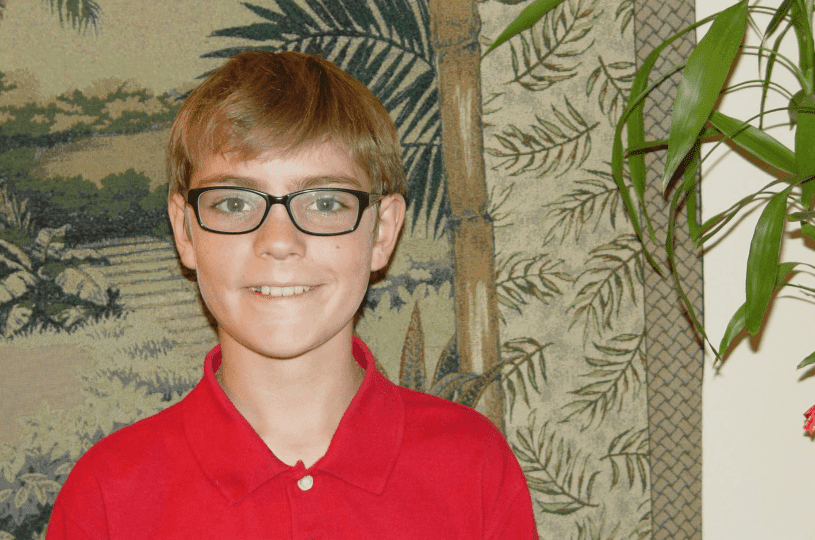 A boy with glasses smiles wearing a red shirt against a leafy backdrop.